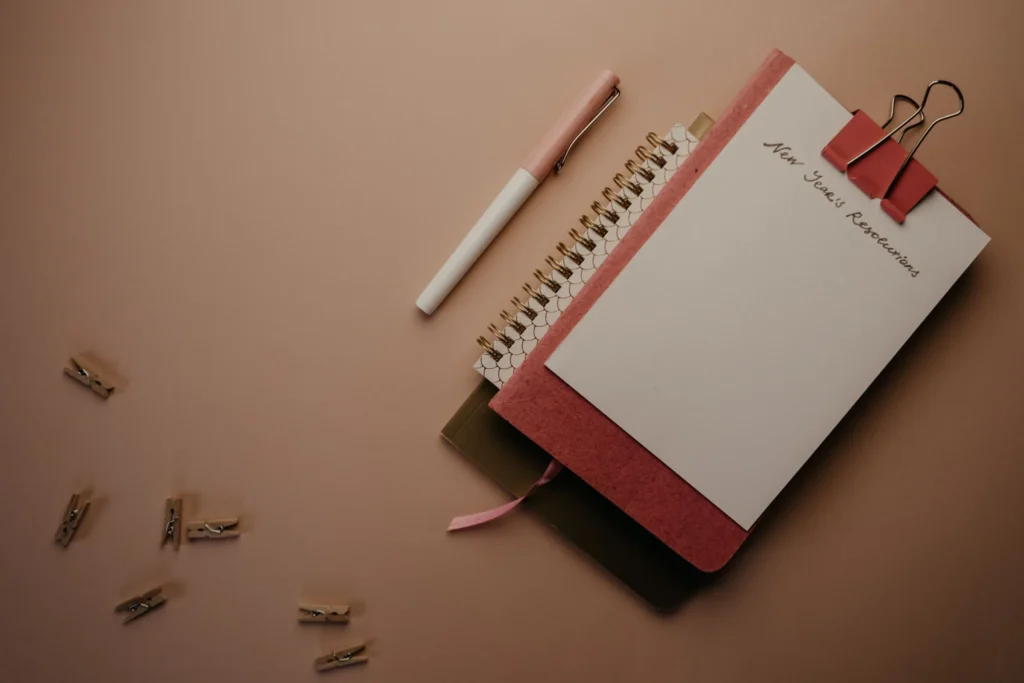 Notebook headed New Year’s Resolutions with a pen and binder clips on a brown surface.