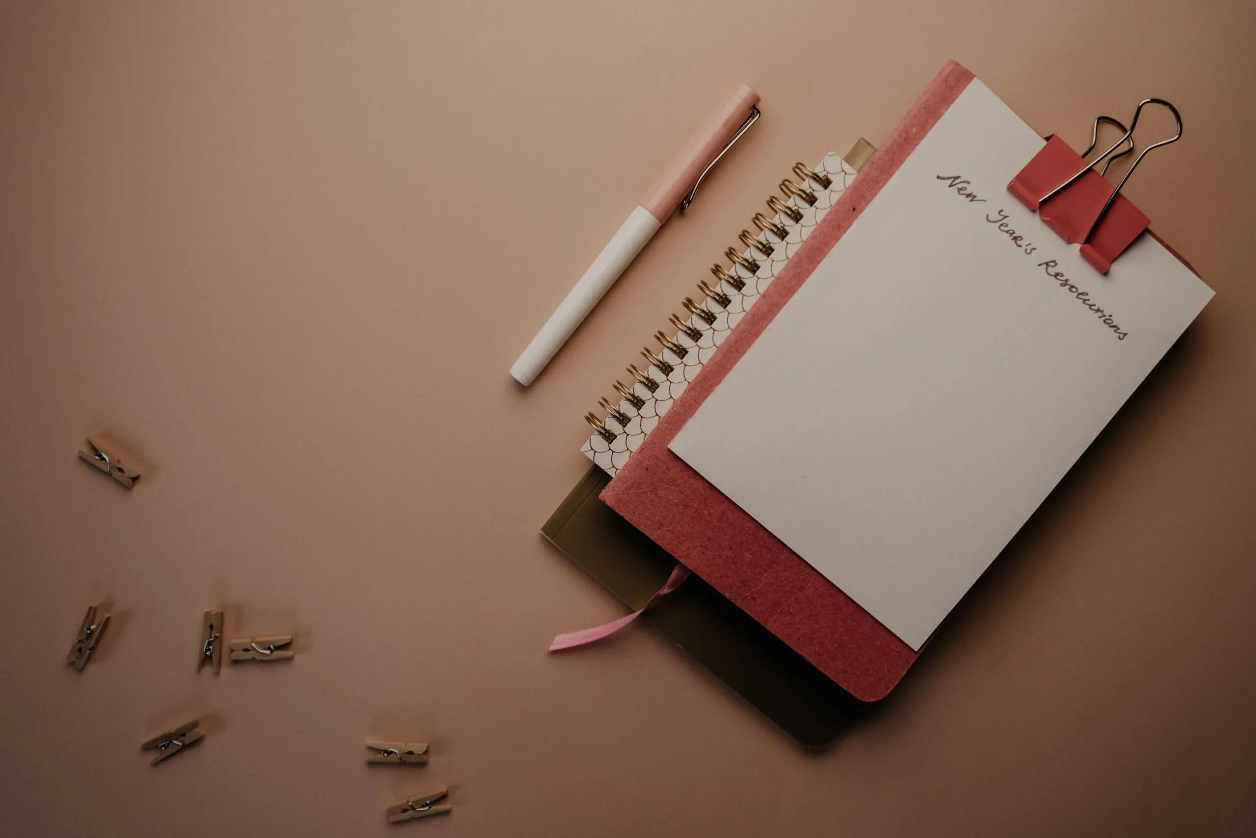 Notebook headed New Year’s Resolutions with a pen and binder clips on a brown surface.