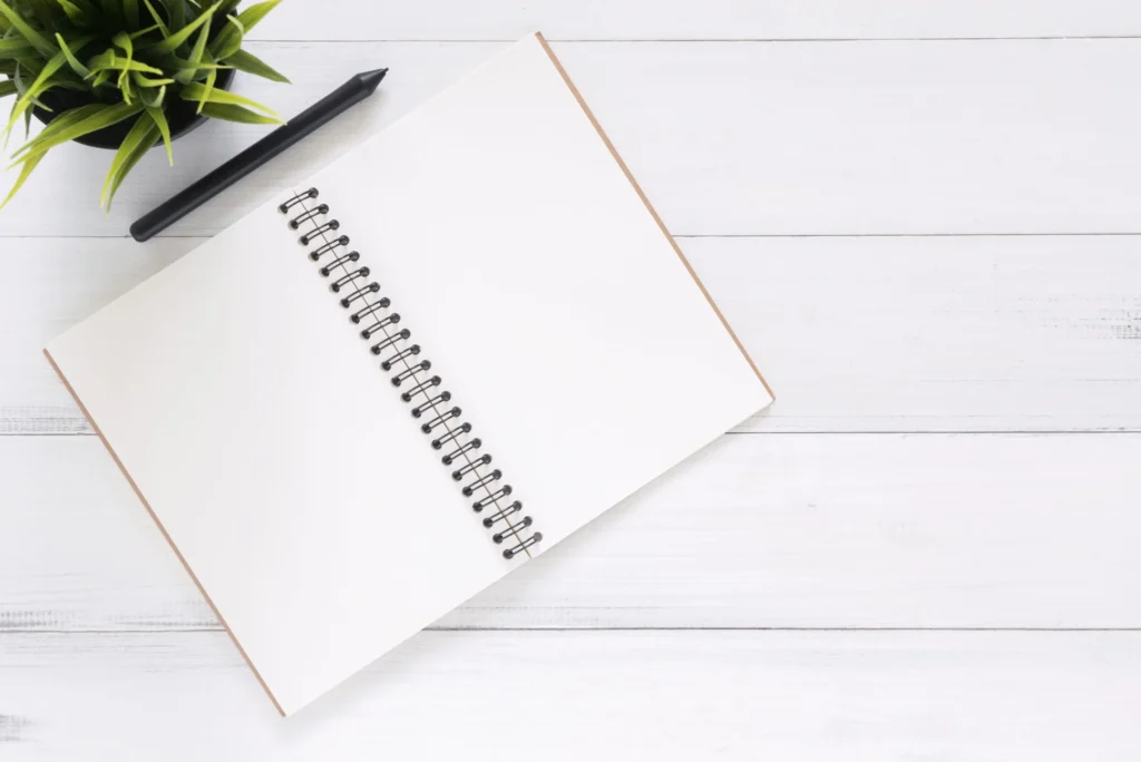 Open blank spiral notebook and black pen on a white desk beside a green plant.