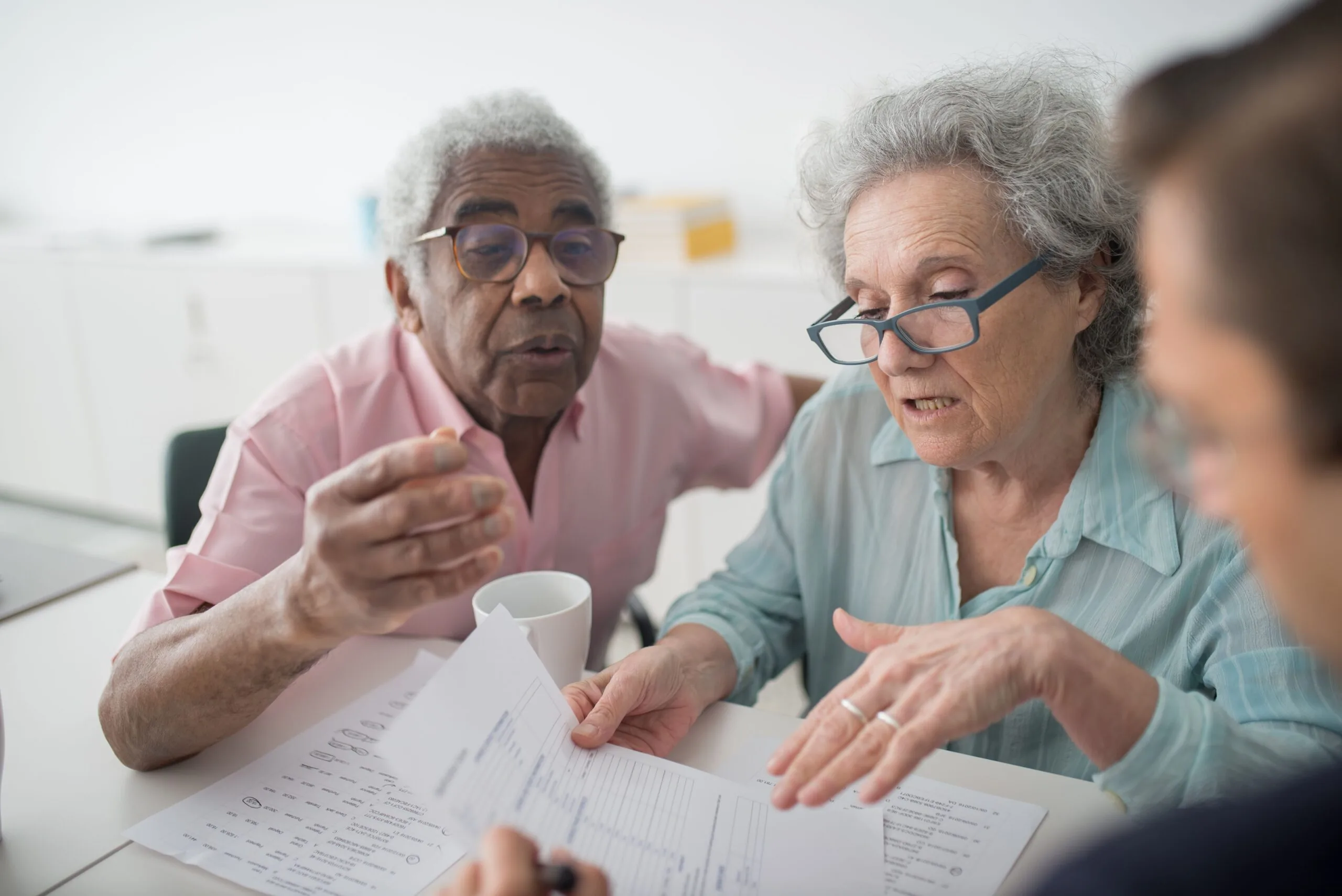 Two older adults discussing documents across a table with another person.